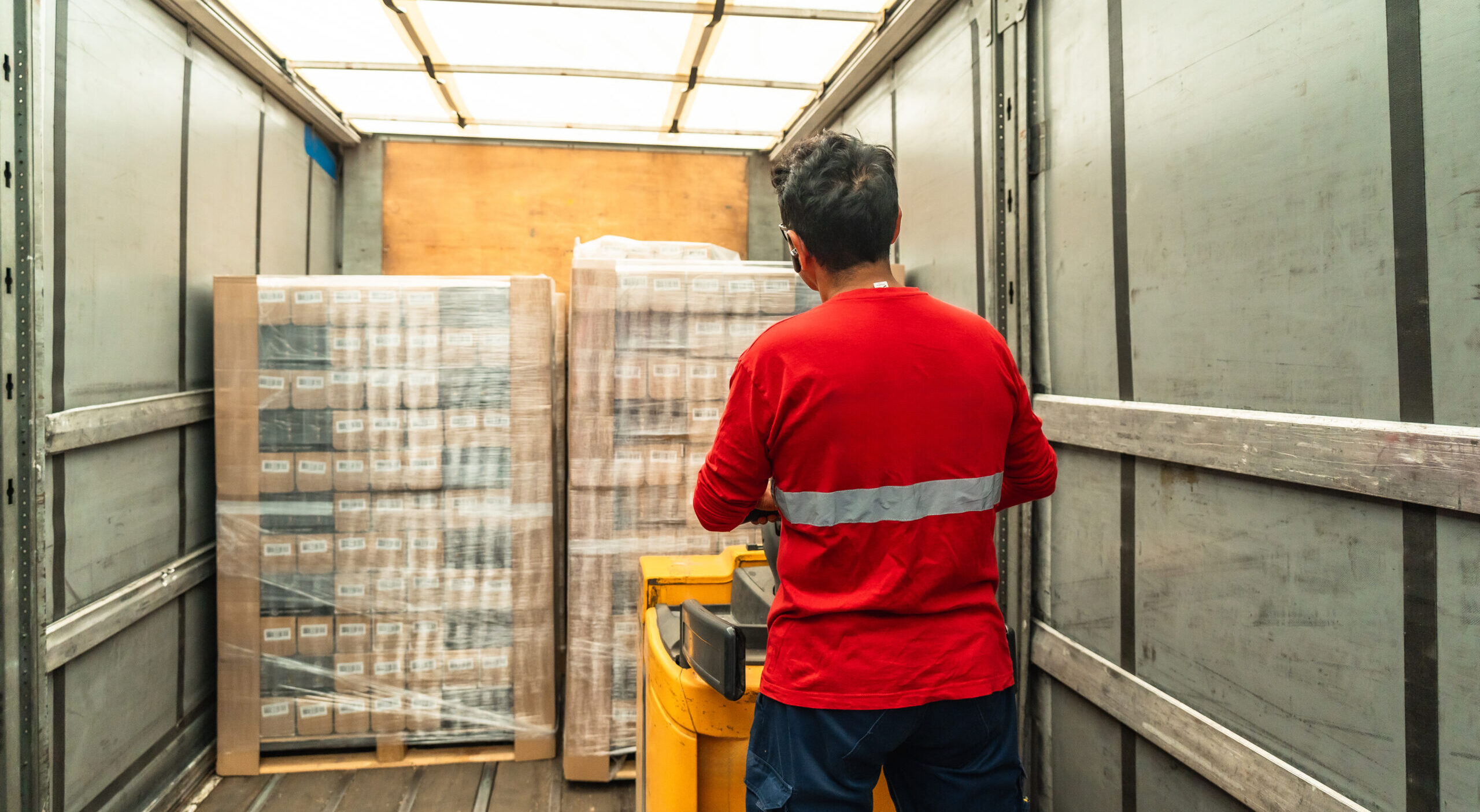 Rear view of a latin male delivery worker loading cardboard cartons on hydraulic hand truck in a warehouse